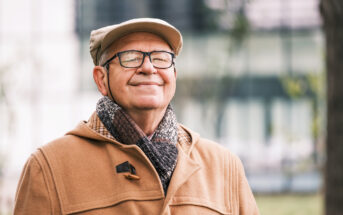 A smiling elderly man is outdoors, wearing a beige coat, a matching flat cap, and a cozy scarf. He has glasses and appears content as he stands in a blurred outdoor setting, possibly a park or garden area.