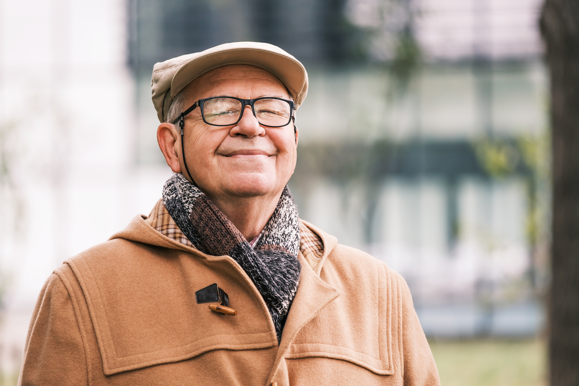 A smiling elderly man is outdoors, wearing a beige coat, a matching flat cap, and a cozy scarf. He has glasses and appears content as he stands in a blurred outdoor setting, possibly a park or garden area.