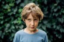 A young boy with messy, light brown hair stands in front of a leafy green background. He is wearing a light blue T-shirt and has a slightly serious or pensive expression on his face.