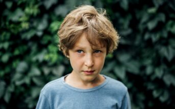 A young boy with messy, light brown hair stands in front of a leafy green background. He is wearing a light blue T-shirt and has a slightly serious or pensive expression on his face.