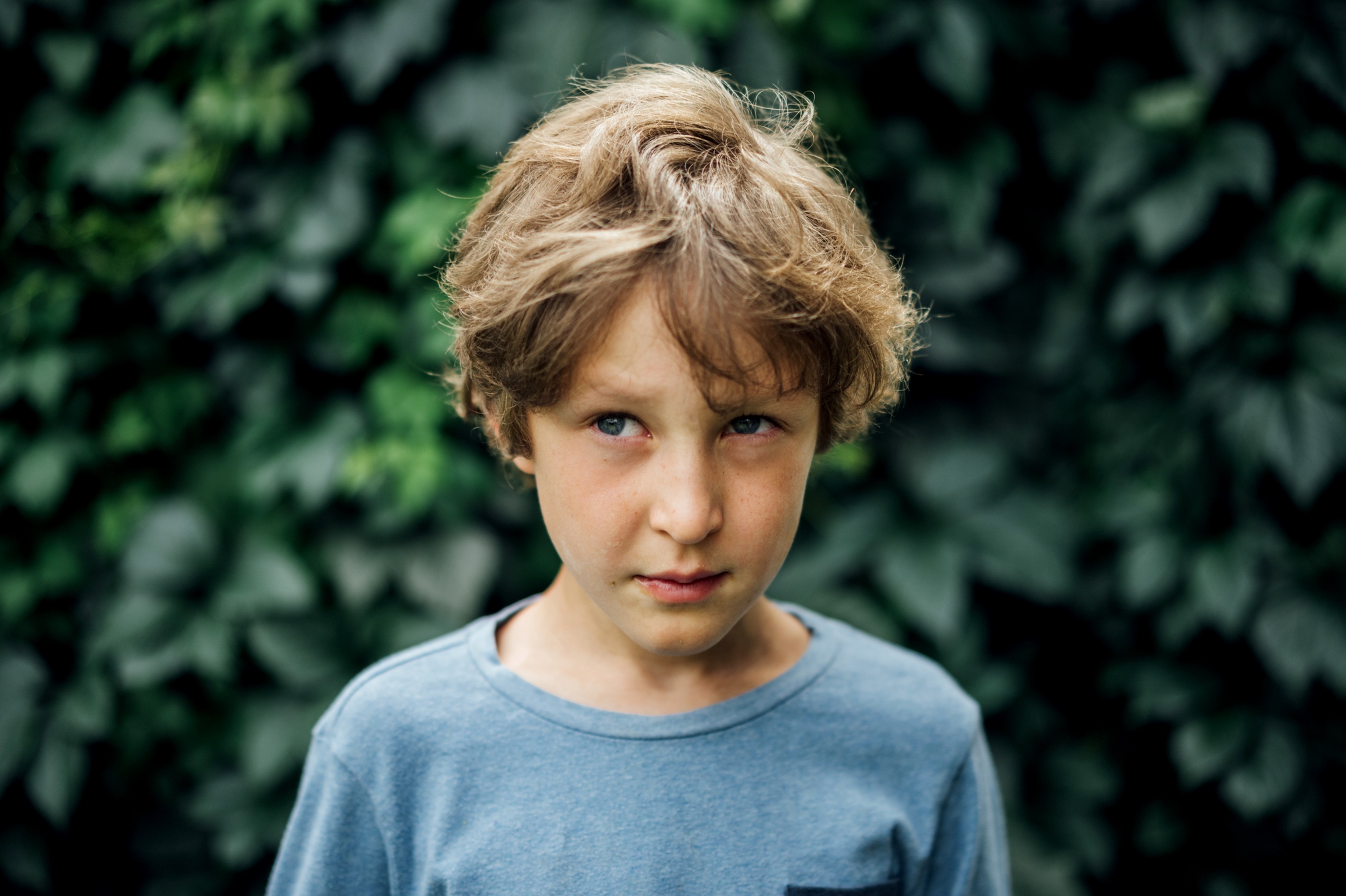 A young boy with messy, light brown hair stands in front of a leafy green background. He is wearing a light blue T-shirt and has a slightly serious or pensive expression on his face.