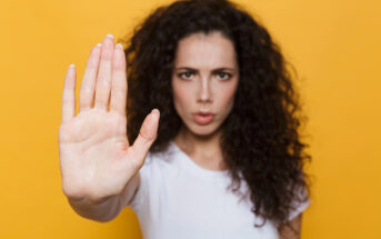 A person with long, curly dark hair and a white shirt holds out their hand towards the camera as if signaling to stop. Their facial expression is serious. The background is a solid bright yellow.