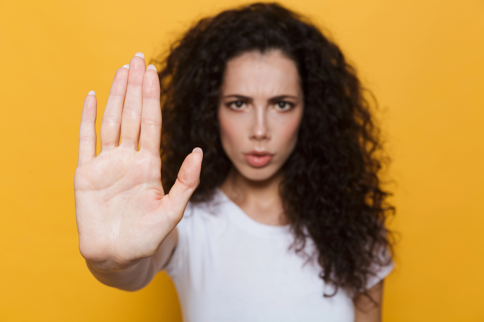 A person with long, curly dark hair and a white shirt holds out their hand towards the camera as if signaling to stop. Their facial expression is serious. The background is a solid bright yellow.