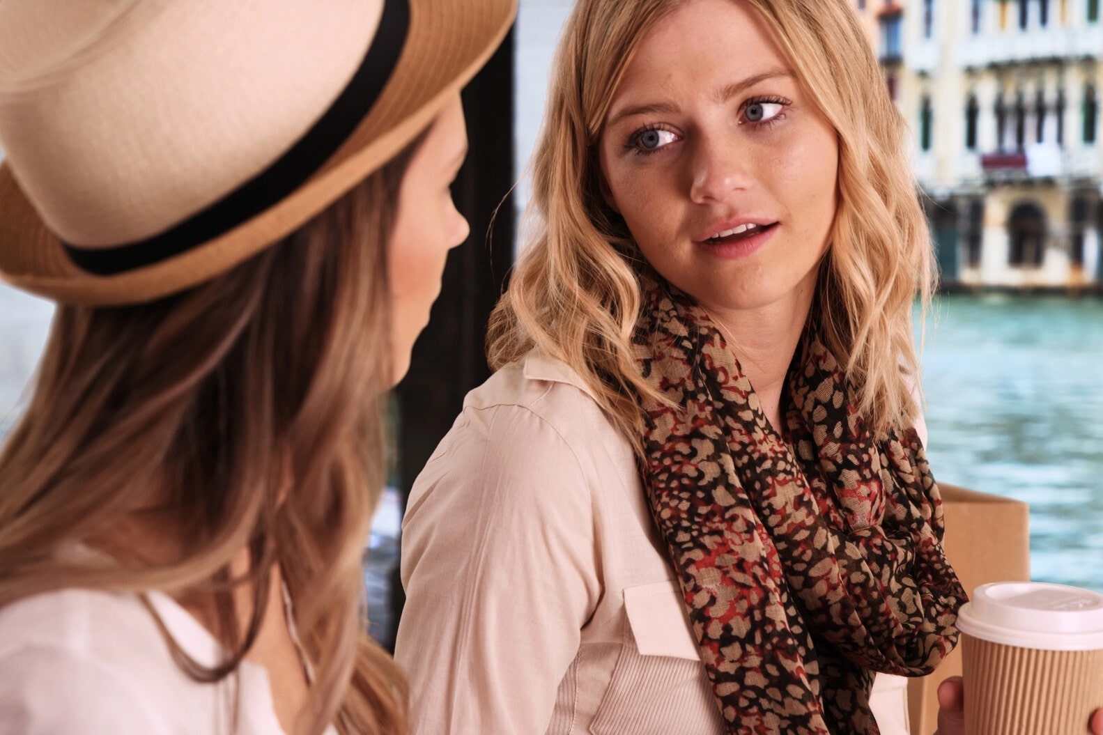 Two women sitting and chatting at an outdoor café with a canal and buildings in the background. One woman, wearing a hat, holds a coffee cup while the other woman, wearing a patterned scarf, looks at her while speaking.