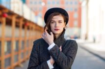 A woman stands in an urban setting with blurred buildings in the background. She wears a dark hat and a grey coat, with one hand adjusting her coat and the other touching her face. She has long, dark hair and a thoughtful expression.