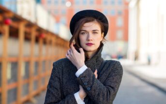 A woman stands in an urban setting with blurred buildings in the background. She wears a dark hat and a grey coat, with one hand adjusting her coat and the other touching her face. She has long, dark hair and a thoughtful expression.