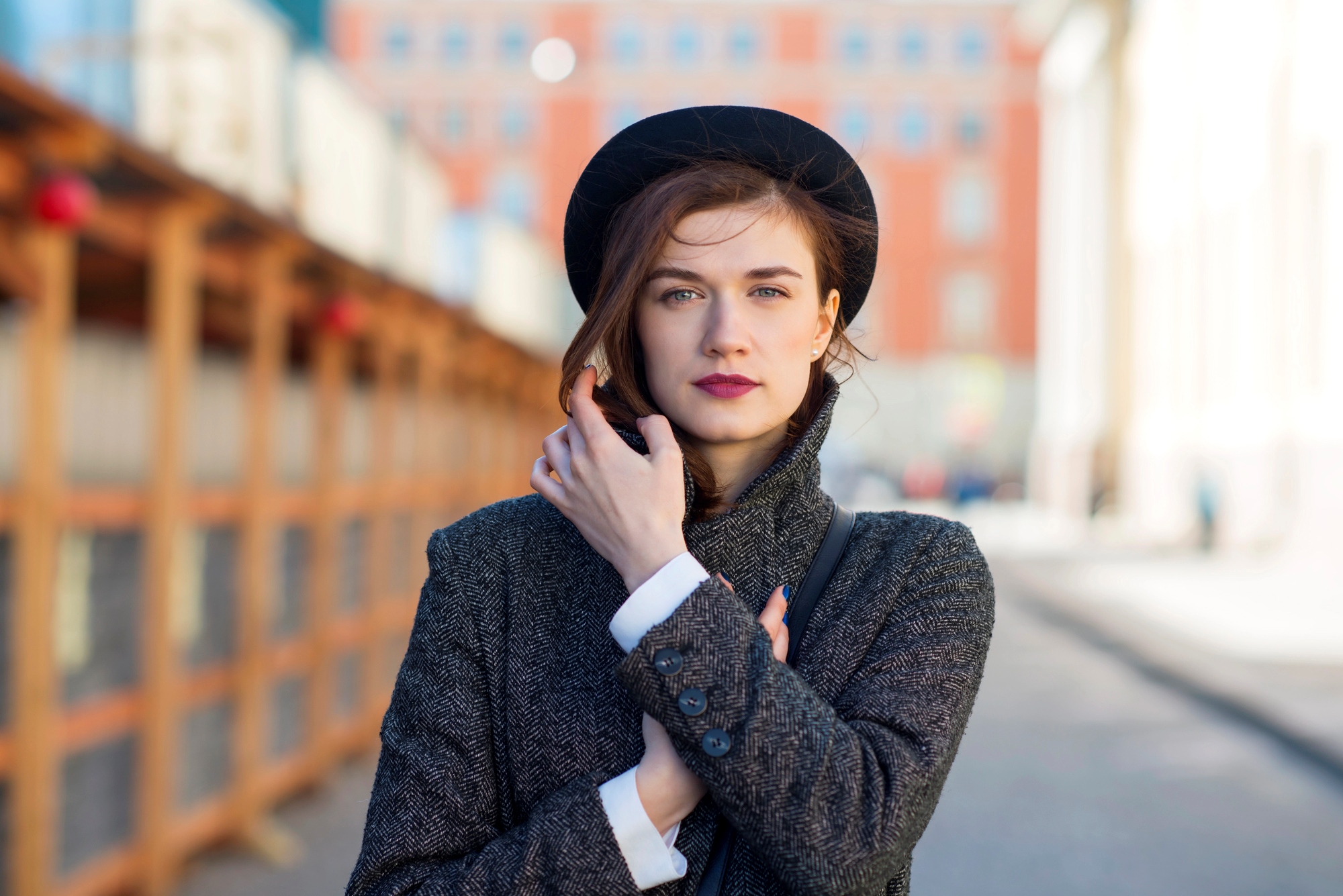 A woman stands in an urban setting with blurred buildings in the background. She wears a dark hat and a grey coat, with one hand adjusting her coat and the other touching her face. She has long, dark hair and a thoughtful expression.