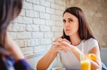 A woman with long dark hair is sitting at a table with a serious expression, hands clasped near her mouth. She wears a white shirt and has a drink with an orange slice on the rim in front of her. She appears to be listening intently to another person.