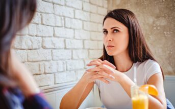 A woman with long dark hair is sitting at a table with a serious expression, hands clasped near her mouth. She wears a white shirt and has a drink with an orange slice on the rim in front of her. She appears to be listening intently to another person.