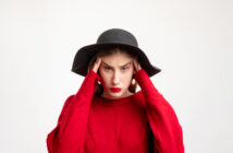 A woman wearing a black wide-brimmed hat and red sweater holds her head with both hands, looking stressed or frustrated. She has long, straight brown hair and is standing against a plain white background.