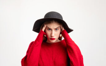 A woman wearing a black wide-brimmed hat and red sweater holds her head with both hands, looking stressed or frustrated. She has long, straight brown hair and is standing against a plain white background.