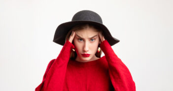 A woman wearing a black wide-brimmed hat and red sweater holds her head with both hands, looking stressed or frustrated. She has long, straight brown hair and is standing against a plain white background.