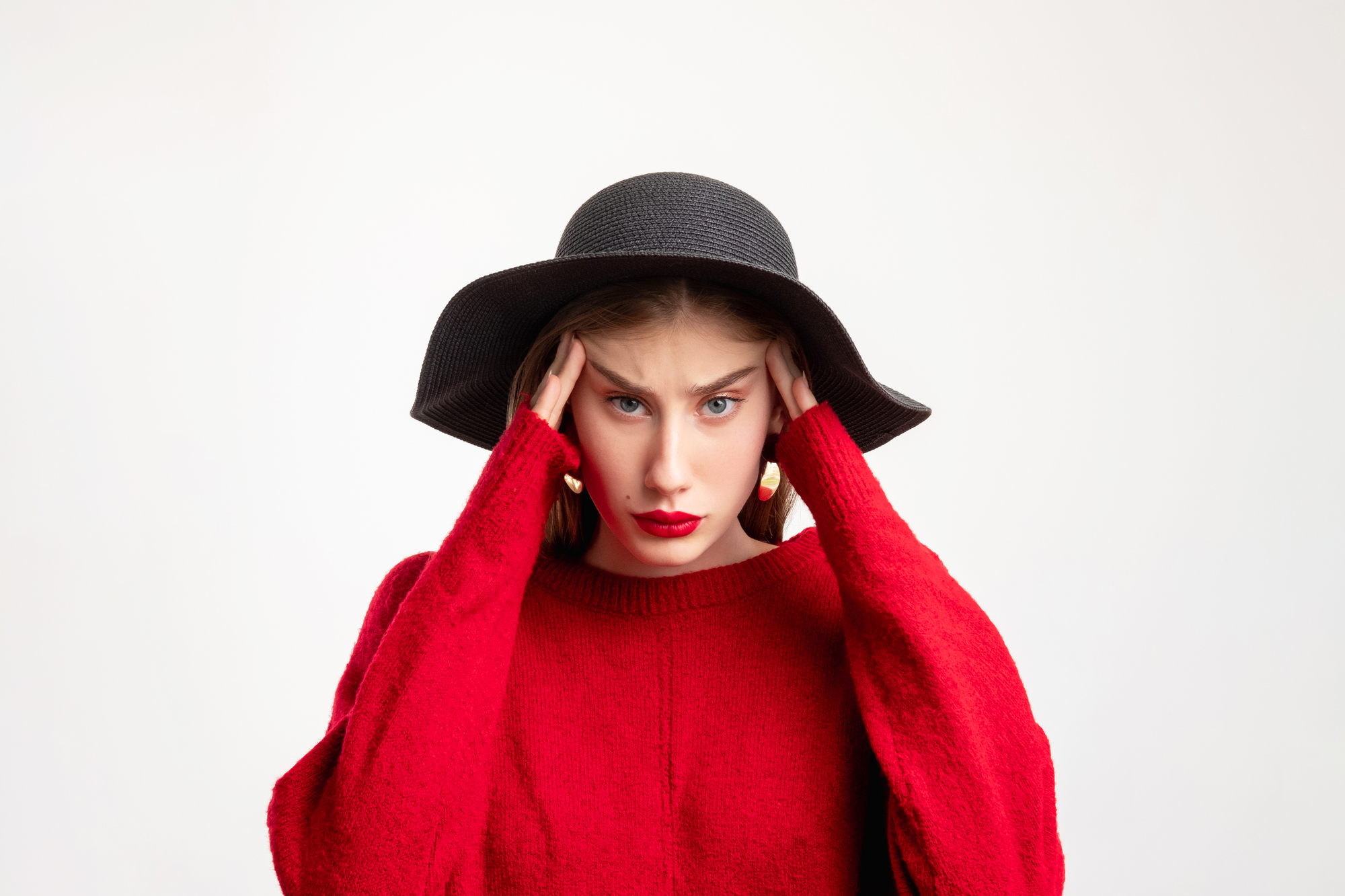 A woman wearing a black wide-brimmed hat and red sweater holds her head with both hands, looking stressed or frustrated. She has long, straight brown hair and is standing against a plain white background.