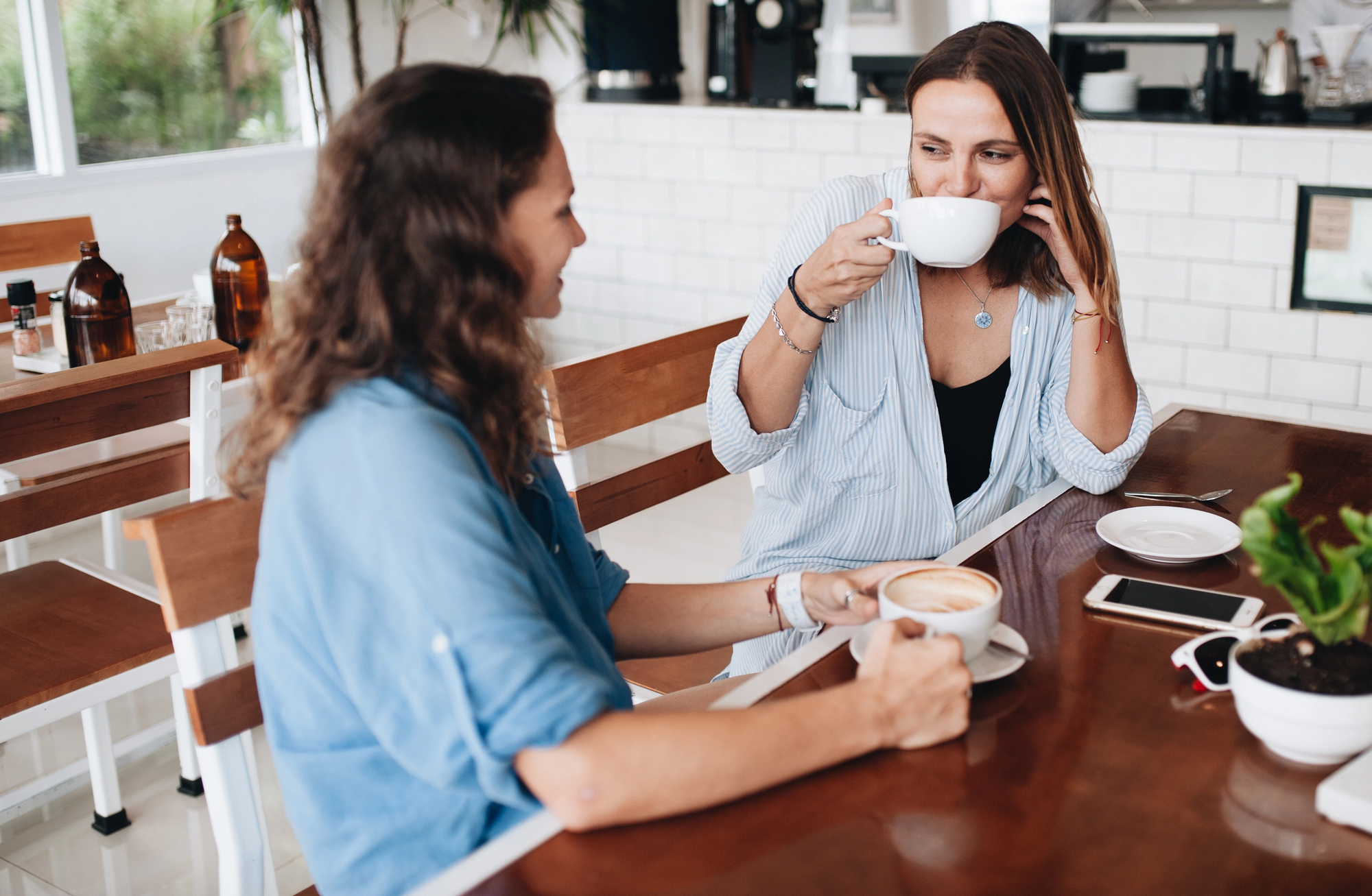 Two individuals sitting at a wooden table in a modern cafe, each holding and sipping from white mugs. Both wear casual attire and appear engaged in conversation. The cafe setting includes white tile decor and visible coffee-making equipment in the background.