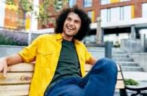 A man with curly hair is sitting on a wooden bench, smiling and looking off to the side. He is wearing a bright yellow shirt over a green t-shirt and blue jeans. The background features modern buildings, greenery, and purple flowers.