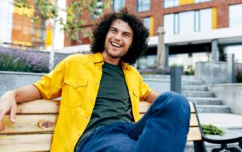 A man with curly hair is sitting on a wooden bench, smiling and looking off to the side. He is wearing a bright yellow shirt over a green t-shirt and blue jeans. The background features modern buildings, greenery, and purple flowers.