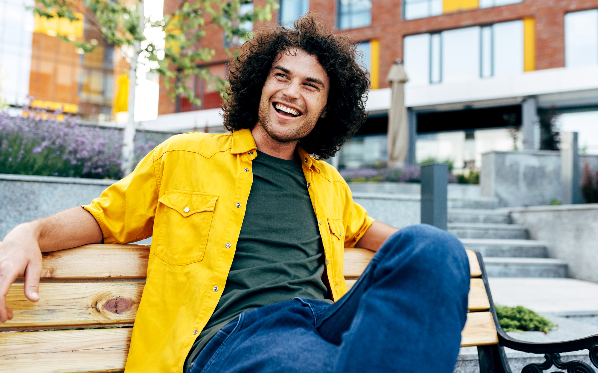 A man with curly hair is sitting on a wooden bench, smiling and looking off to the side. He is wearing a bright yellow shirt over a green t-shirt and blue jeans. The background features modern buildings, greenery, and purple flowers.