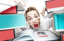 A person with red lipstick and white-framed glasses looks stressed and overwhelmed while peering through a large stack of binders. A calculator and a notebook are placed in front of them on a desk.