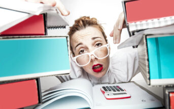 A person with red lipstick and white-framed glasses looks stressed and overwhelmed while peering through a large stack of binders. A calculator and a notebook are placed in front of them on a desk.