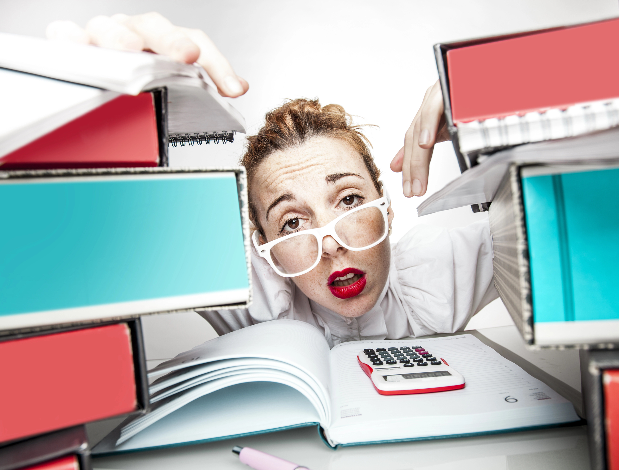 A person with red lipstick and white-framed glasses looks stressed and overwhelmed while peering through a large stack of binders. A calculator and a notebook are placed in front of them on a desk.