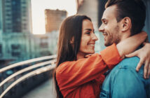 A woman in an orange jacket embraces and smiles at a man in a blue jacket who is smiling back at her. They are standing close on a city bridge or walkway with blurred buildings and a setting sun in the background.