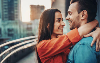 A woman in an orange jacket embraces and smiles at a man in a blue jacket who is smiling back at her. They are standing close on a city bridge or walkway with blurred buildings and a setting sun in the background.