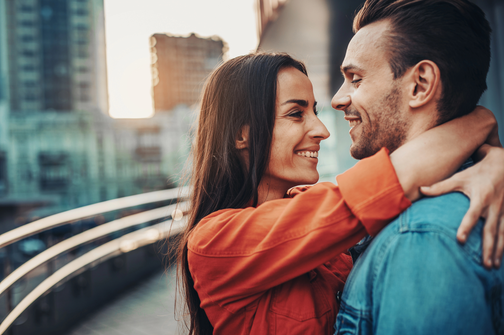 A woman in an orange jacket embraces and smiles at a man in a blue jacket who is smiling back at her. They are standing close on a city bridge or walkway with blurred buildings and a setting sun in the background.