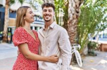 A smiling couple stands outside in a sunny area with palm trees and a parked bicycle in the background. The woman wears a red floral dress, and the man is dressed in a light beige shirt. They are embracing and looking at each other.