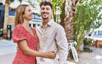 A smiling couple stands outside in a sunny area with palm trees and a parked bicycle in the background. The woman wears a red floral dress, and the man is dressed in a light beige shirt. They are embracing and looking at each other.