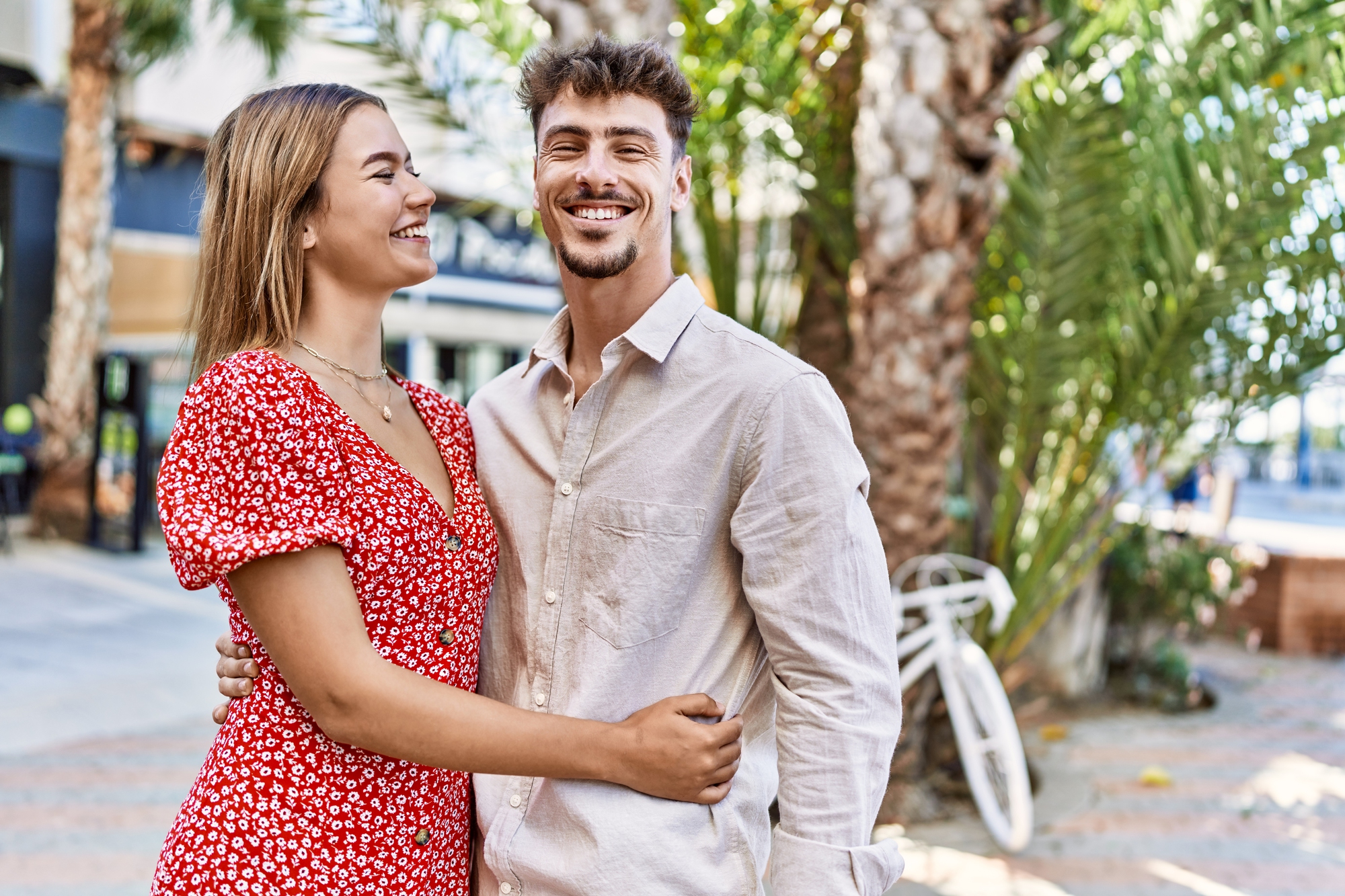 A smiling couple stands outside in a sunny area with palm trees and a parked bicycle in the background. The woman wears a red floral dress, and the man is dressed in a light beige shirt. They are embracing and looking at each other.