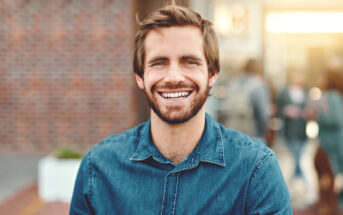 A bearded man with tousled brown hair smiles warmly at the camera. He is wearing a blue denim shirt and stands in front of a blurred outdoor background with brick and people in soft focus.