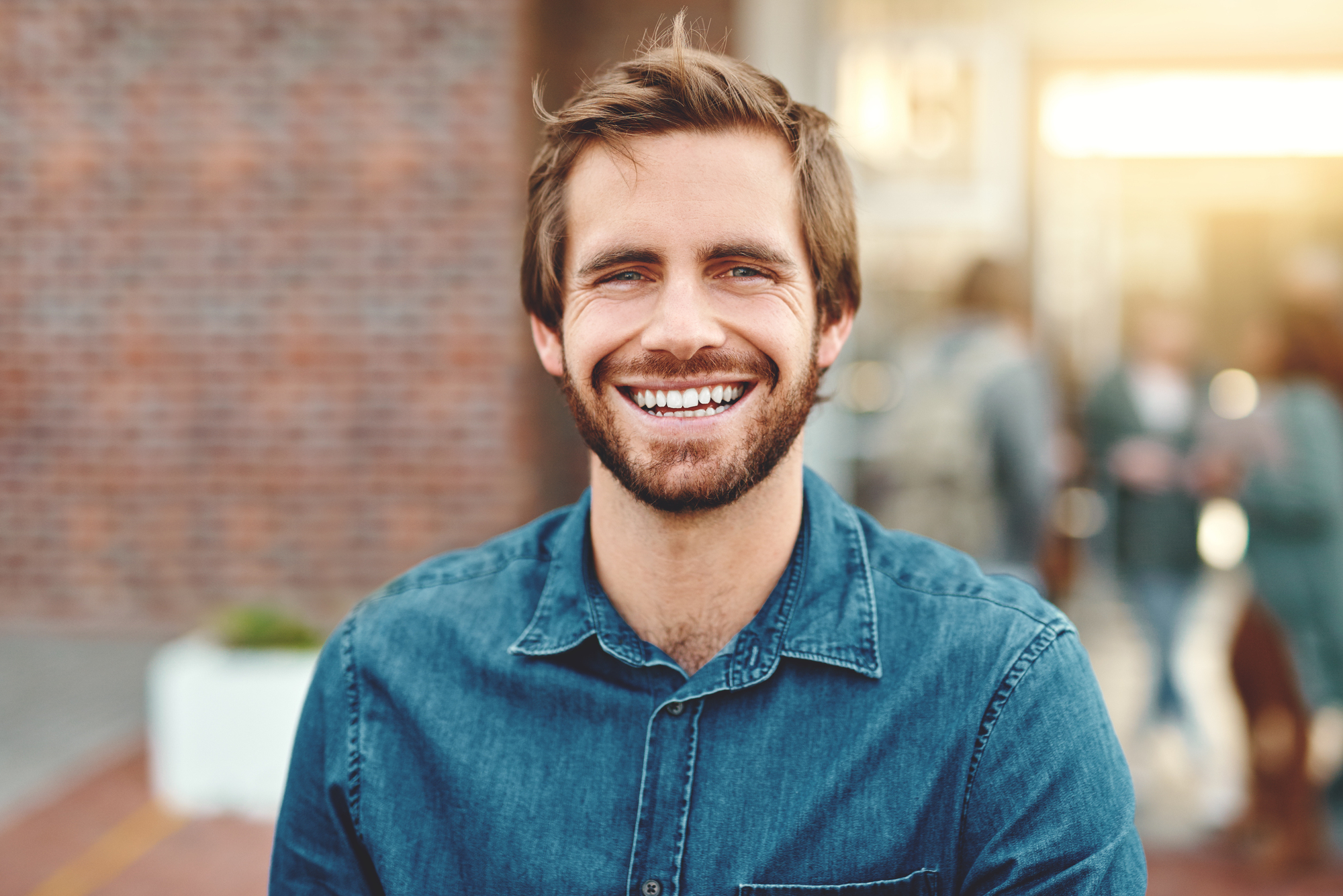 A bearded man with tousled brown hair smiles warmly at the camera. He is wearing a blue denim shirt and stands in front of a blurred outdoor background with brick and people in soft focus.