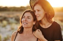 Two women standing outdoors at sunset in a natural setting. The woman on the right, wearing a black shirt and brimmed hat, rests her arm affectionately on the shoulder of the woman on the left, who is wearing a tank top. Both gaze into the distance with serene expressions.