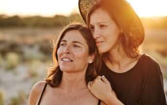 Two women standing outdoors at sunset in a natural setting. The woman on the right, wearing a black shirt and brimmed hat, rests her arm affectionately on the shoulder of the woman on the left, who is wearing a tank top. Both gaze into the distance with serene expressions.