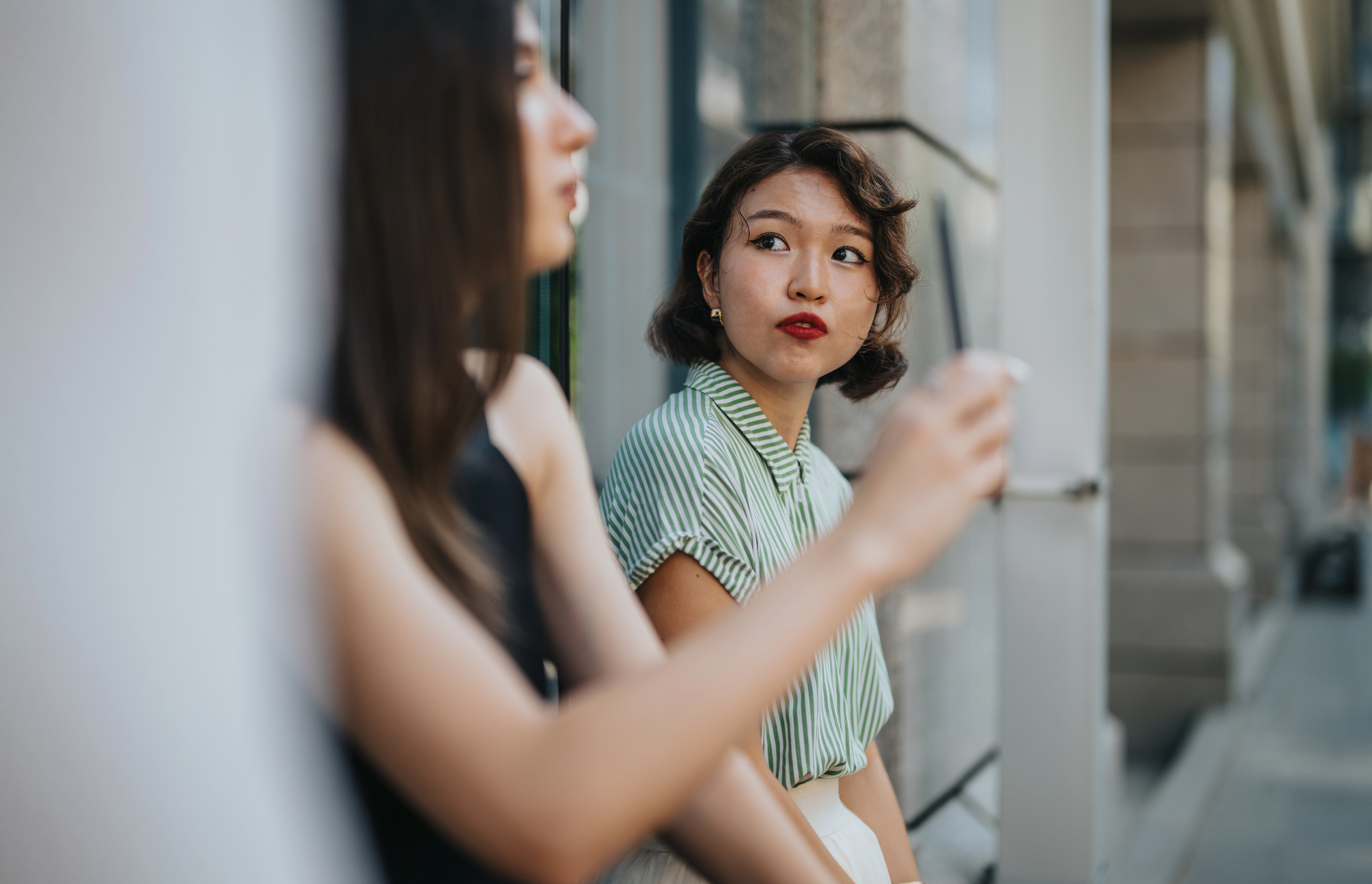 Two women are sitting outside a building. The woman on the left is blurred and facing away, wearing a dark top. The woman on the right is in focus, wearing a striped shirt, and holding a cup, looking thoughtfully at the other woman. The background shows a city street.