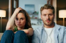 A woman and a man sit closely on a couch, looking intently at the camera. The woman has her hand on her head, while the man has a neutral expression. Both have light skin and brown hair. The room behind them is softly lit with lamps and a blurred shelf in the background.