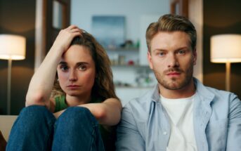 A woman and a man sit closely on a couch, looking intently at the camera. The woman has her hand on her head, while the man has a neutral expression. Both have light skin and brown hair. The room behind them is softly lit with lamps and a blurred shelf in the background.