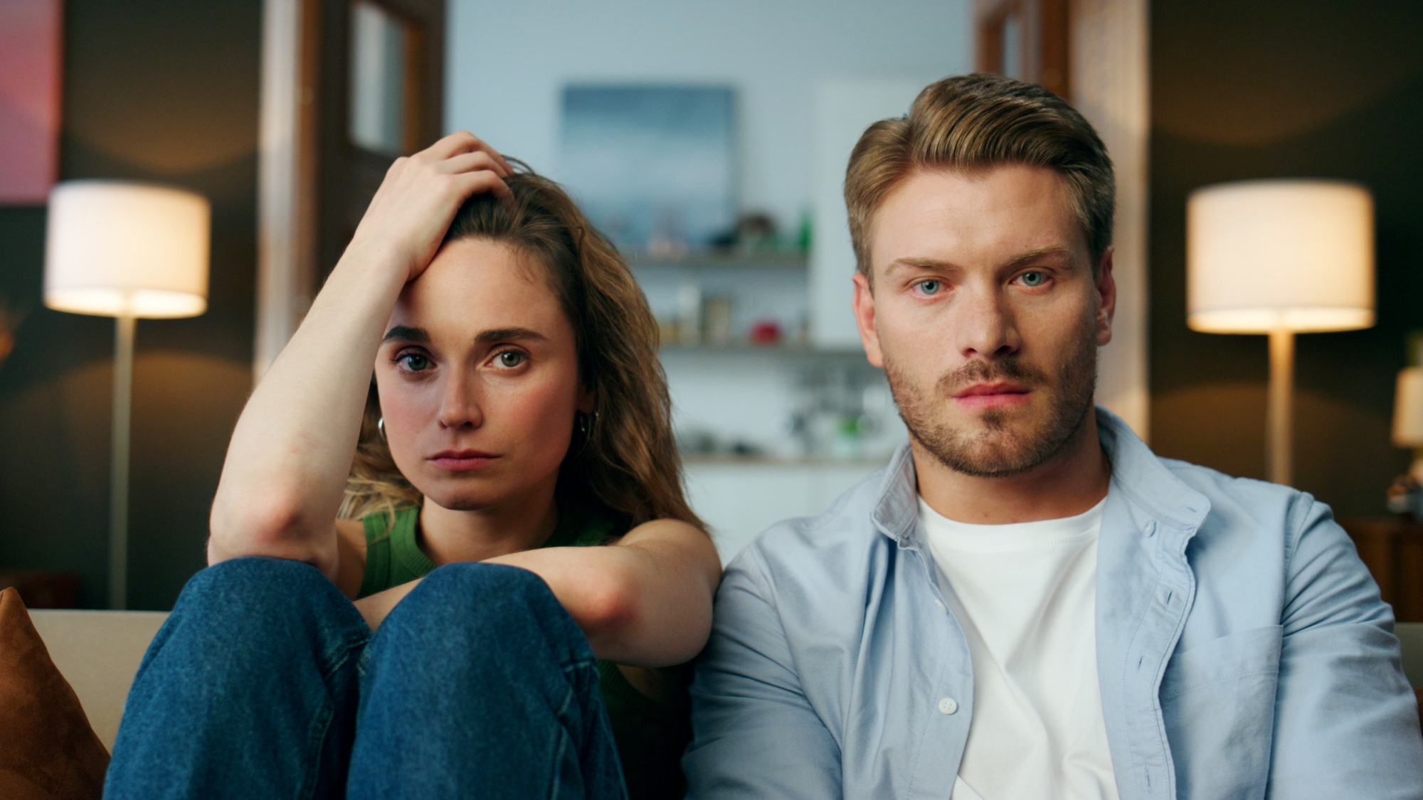 A woman and a man sit closely on a couch, looking intently at the camera. The woman has her hand on her head, while the man has a neutral expression. Both have light skin and brown hair. The room behind them is softly lit with lamps and a blurred shelf in the background.