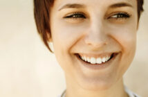 Close-up of a person with short brown hair smiling broadly. Their eyes are slightly squinted, expressing happiness and warmth. The background is blurred, emphasizing the person's face and joyful expression.