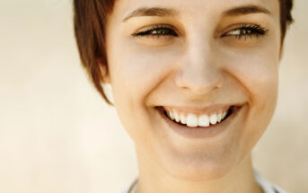 Close-up of a person with short brown hair smiling broadly. Their eyes are slightly squinted, expressing happiness and warmth. The background is blurred, emphasizing the person's face and joyful expression.