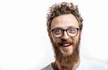 A person with curly hair, a full beard, and glasses is smiling widely. They are wearing a white shirt and are positioned against a plain white background. The image captures a close-up of their face, expressing a joyful and enthusiastic demeanor.