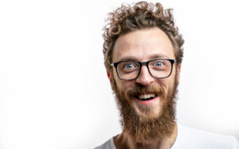 A person with curly hair, a full beard, and glasses is smiling widely. They are wearing a white shirt and are positioned against a plain white background. The image captures a close-up of their face, expressing a joyful and enthusiastic demeanor.