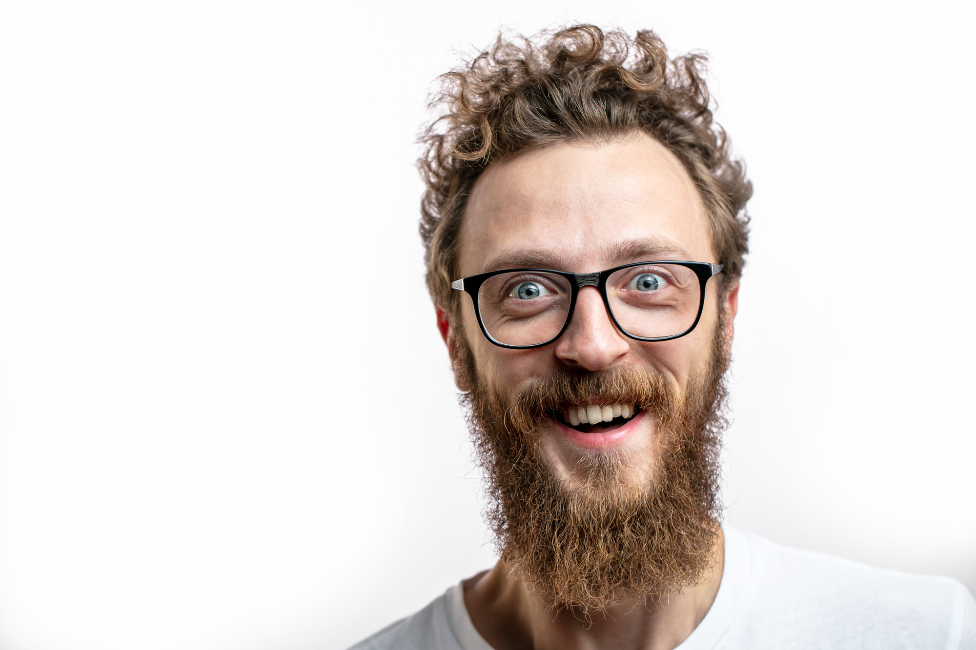 A person with curly hair, a full beard, and glasses is smiling widely. They are wearing a white shirt and are positioned against a plain white background. The image captures a close-up of their face, expressing a joyful and enthusiastic demeanor.