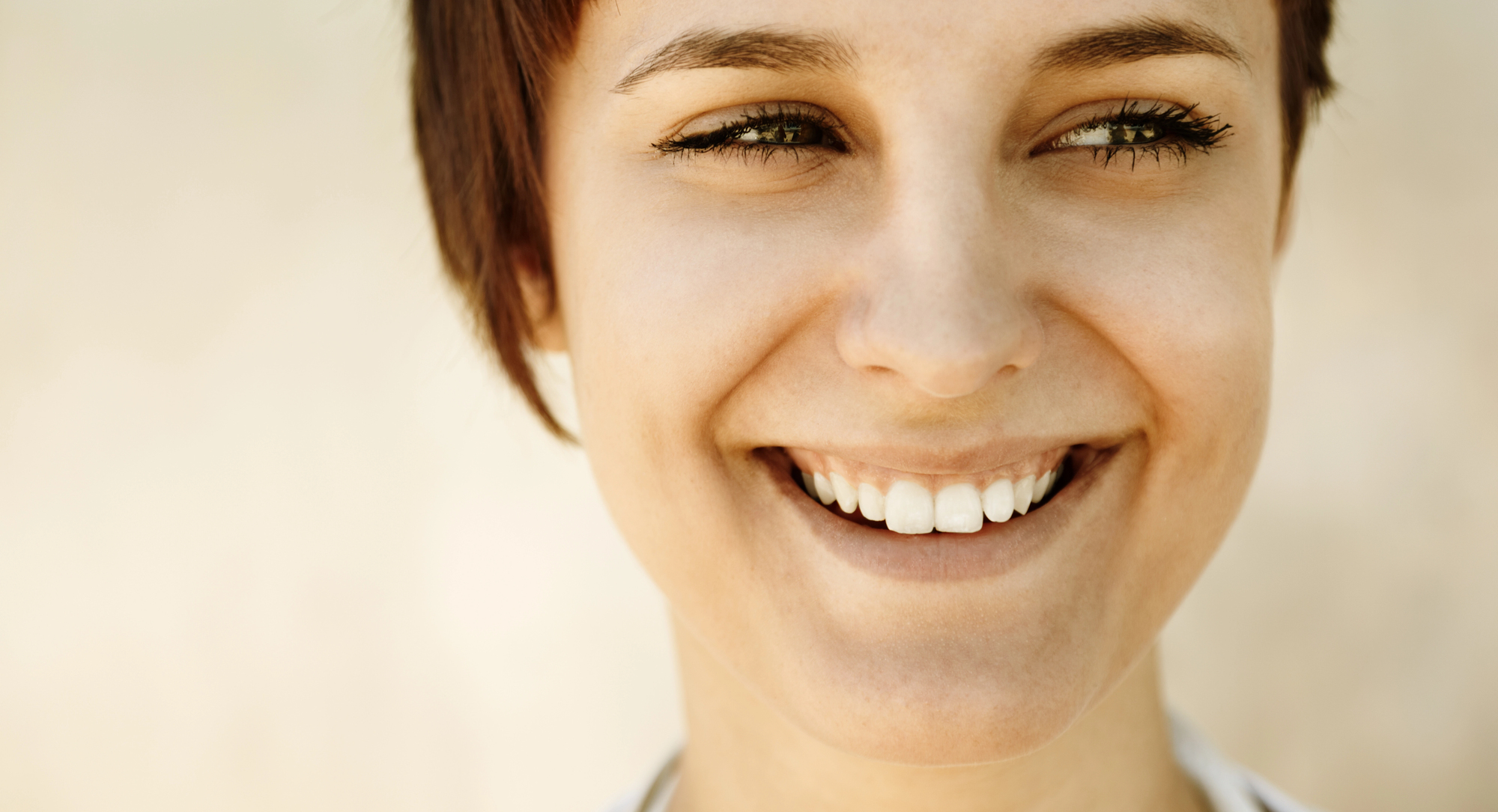 Close-up of a person with short brown hair smiling broadly. Their eyes are slightly squinted, expressing happiness and warmth. The background is blurred, emphasizing the person's face and joyful expression.