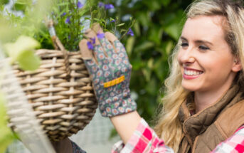 A smiling woman wearing patterned gloves and a plaid shirt tends to a hanging basket of purple flowers in a garden. She has long, wavy blonde hair and appears to be enjoying her gardening activity.