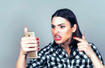 A woman with medium-length dark hair and wearing a black and white checkered shirt angrily points at a smartphone in her other hand. Her facial expression shows frustration and anger as she looks at the phone's screen.