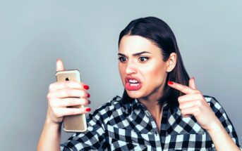 A woman with medium-length dark hair and wearing a black and white checkered shirt angrily points at a smartphone in her other hand. Her facial expression shows frustration and anger as she looks at the phone's screen.