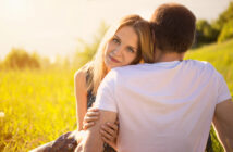 A woman with long blonde hair rests her head on a man's shoulder while sitting on a grassy field. The man, wearing a white T-shirt, faces away from the camera. Warm sunlight illuminates the scene, evoking a serene and intimate moment.