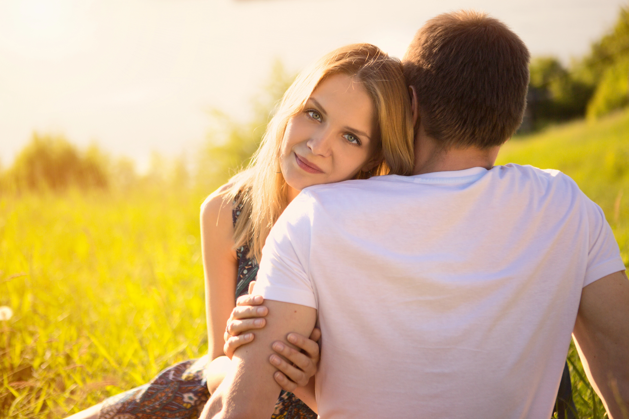 A woman with long blonde hair rests her head on a man's shoulder while sitting on a grassy field. The man, wearing a white T-shirt, faces away from the camera. Warm sunlight illuminates the scene, evoking a serene and intimate moment.