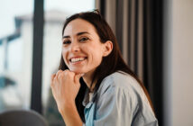 A woman with long dark hair is smiling warmly while resting her chin on her hand. She is wearing a light blue denim shirt and has a pair of glasses perched on her head. The background is softly blurred, showing a window with curtains and a glimpse of an outdoor scene.